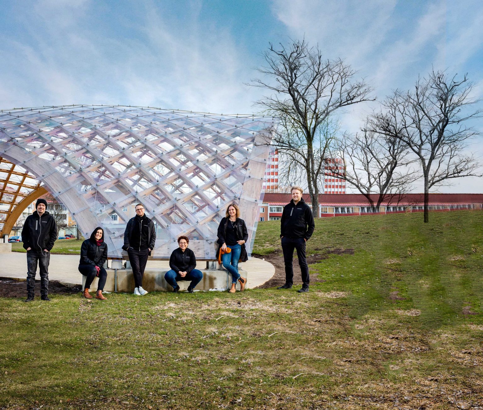 Sex personer står uppställda framför ett stort konstverk utomhus. De ler och tittar in i kameran.